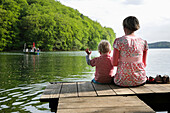 Mutter und Sohn sitzen auf einem Steg am Schmalen Luzin, Luzinfähre im Hintergrund, Naturpark Feldberger Seenlandschaft, Mecklenburg-Vorpommern, Deutschland