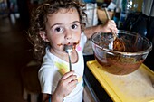 Young girl licking chocolate spoons in a kitchen