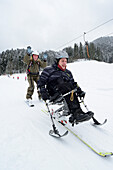 Mann mit Gehbehinderung fährt mit Begleiterin auf Piste Ski, Söll, Kitzbüheler Alpen, Tirol, Österreich