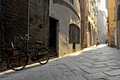 Fahrrad und Radfahrerin im morgendlichen Streiflicht in einer Gasse von Lucca, Toskana, Italien