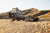 Himbafrauen und Touristin sitzen neben einem Geländewagen, Hartmanntal, Kaokoland, Kunene, Namibia