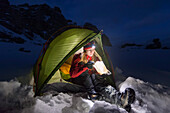 Young woman with headlamp eating in front of a tent in snow, the Dolomites, Belluno, Veneto, Italy