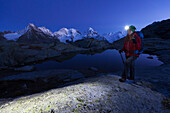 Female hiker with headlamp passing a mountain lake above the Roseg valley, Piz Morteratsch, Piz Bernina and Piz Roseg in background, Engadin, Canton of Grisons, Switzerland