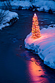 Lit Christmas Tree On The Bank Of A Stream During Winter In Southcentral Alaska