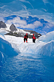 Two Hikers Inpect A Melting Ice Cave On Mendenhall Glacier Near Juneau. Summer In Southeast Alaska.