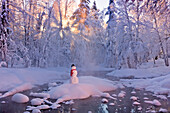 Snowman standing on a small island in the middle of a stream with sunrays shining through fog and hoar frosted trees in the background, Russian Jack Springs Park, Anchorage, Southcentral Alaska, Winter. Digitally enhanced.