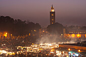 Looking across food stalls in Dejmaa el Fna at dusk with minaret of Koutoubia Mosque behind, Marrakesh, Morocco