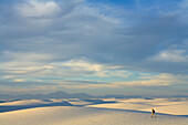 Man hiker wearing shorts, a hat and a backpack, 52, walking alone in a big sprawling landscape of sand and distant mountains, across white sand dunes, evening, autumn, White Sands National Monument, New Mexico