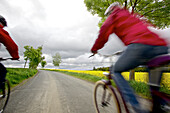 Two cyclists with electric bicycles passing blooming canola field, Tanna, Thuringia, Germany