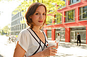 young woman with coffee cup in front of modern building, Munich, Bavaria, Germany
