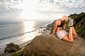 A young woman performing yoga on cliffs above the beach at sunset Malibu, California, USA