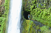 A woman running along trail blasted into a hill behind a waterfall on the Pacific Crest Trail, Oregon Eagle Creek, Oregon, USA