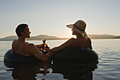 A young couple relax with a cold beverage while floating in a lake at sunset in inner tubes Sandpoint, Idaho, USA
