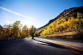 A young woman longboards down a smooth country road through the mountain peaks and gold forests Aspen, Colorado, USA
