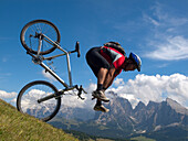 A mountain biker tumbles over his bike while riding downhill a grassy slope at Seiser Alm, with rock cliffs in the background Val Gardena, Dolomites, Italy