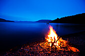 A beautiful camp fire burns at dusk against a deep blue lake with a wakeboard boat in the background in Idaho Sandpoint, Idaho, USA