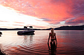 A beautiful young woman standing in a lake at sunset next to a wakeboard boat in Idaho., Sandpoint, Idaho, USA