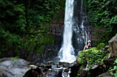 A beautiful young woman sitting in a lush jungle next to a waterfall in Bali, Indonesia., Lovina, Bali, Indonesia