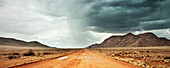 sandige rote Straße mit starkem Regen und Gewitter am Horizont, Tiras Gebirge, Tirasberge, Namib Naukluft Park, Namibia, Afrika