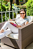 Frau sitzt auf einem Balkon mit Loungemöbeln und blättert in einer Zeitschrift, Hamburg, Deutschland