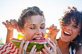Young woman eating watermelon as friend pulls her ears