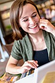 A smiling 18 year old girl in a library reading from a computer screen.