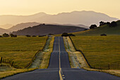Golden sunrise light over long straight two lane country road and rolling hills in Spring, Santa Ynez Valley, California.