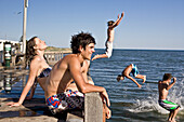 Young Couple Sitting on Edge of Pier With Group of Boys Jumping in Water