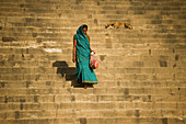 Woman with a water green cloth walks down the stairs of a ghat while a dog sleeps in Varanasi, India