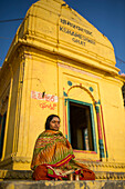 Woman wearing typical indian dress doing yoga and praying outside a yellow temple in the Kshameshwar ghat under a blue sky in Varanasi, India