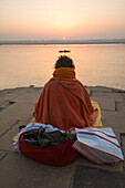 Old man praying at sunrise from the banks of the Ganges river seen from behind while a boat is passing and the sun is rising in the background, Varanasi, India