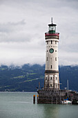 'Old Lighthouse On A Pier Of A Lake With Cloud Covered Mountains In The Background; Lindau, Germany'