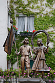 'Bronze Statues Of A Boy And Girl With An Ivy Covered Building And Flowers In The Background; Lindau, Germany'
