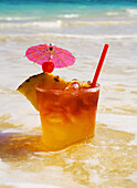 A Mai Tai Garnished With Pinapple And A Cherry, Sitting In Shallow Water On The Beach.