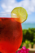 A Bright Red Tropical Cocktail Garnished With Lime In Outdoor Setting.