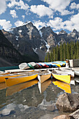 'Canoes Along A Dock Reflecting Off The Lake With Mountains Clouds And Blue Sky; Alberta, Canada'