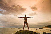 Eine Touristin im Bikini hebt die Arme und macht Yoga am Strand einer tropischen Insel bei Sonnenuntergang; Koh Lanta, Thailand