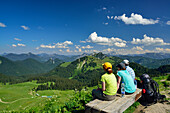 Wanderer sitzen auf einer Bank am Gipfel des Seekarkreuz mit Blick auf Rauhalm, Buchstein, Roßstein und Guffert, Bayerische Voralpen, Oberbayern, Bayern, Deutschland