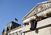 A young man sitting on a wall in front of the Reichstag German parliamentary building reading a newspaper, Berlin, Germany, Europe