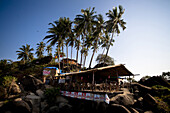 An open air bamboo restaurant with palm trees, perched on a rock outcrop over the sea, in in the beach village of Palolem, in Goa, India.