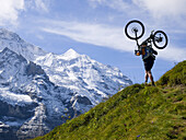 A mountain biker is carrying his bike on a steep trail above Grindelwald. In the background the Silberhorn and Jungfrau mountains.