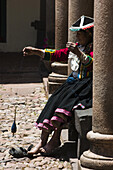 An elderly woman in traditional clothing hand spins alpaca wool under the columns of a stone arcade in Cusco, Peru.