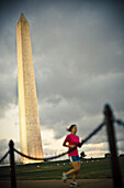 A woman runs by the Washington Monument during sunset with chain fence in the foreground.