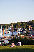 MAINE, USA. A couple sits on a grassy hilltop overlooking the harbor below.