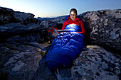 Susann Scheller is sitting against a rock in her sleeping bag while reading a book with a headlamp. Table Mountain National Park. South Africa.