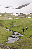 Two skiers hiking up a grassy slope next to a stream on Umnak island in the Aleutian Islands.