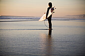 Surfer stands on the beach and looks out at the sunset in Oceanside, CA.