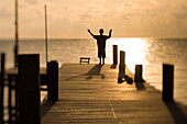 Nico Steele, age 12, holds his arms high while silhouetted against the morning sun on  a wooden dock on Ambergris Caye, Belize.