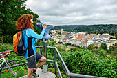 Female cyclist looking through binoculars over Inn river to Wasserburg, Upper Bavaria, Germany