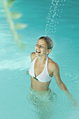 Young woman in pool, standing under stream of water, eyes closed